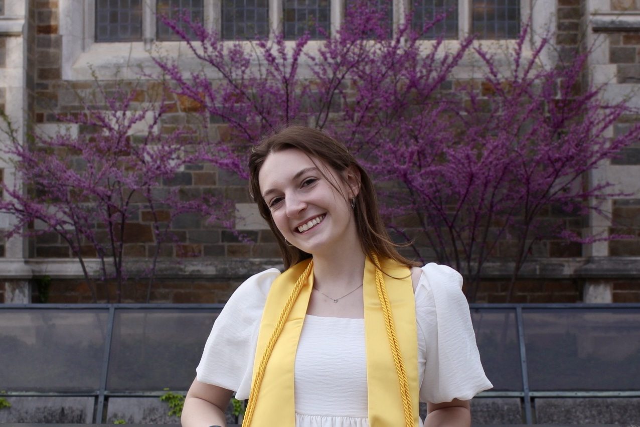 Maya Wolock in a white shirt and yellow stole in front of purple trees.